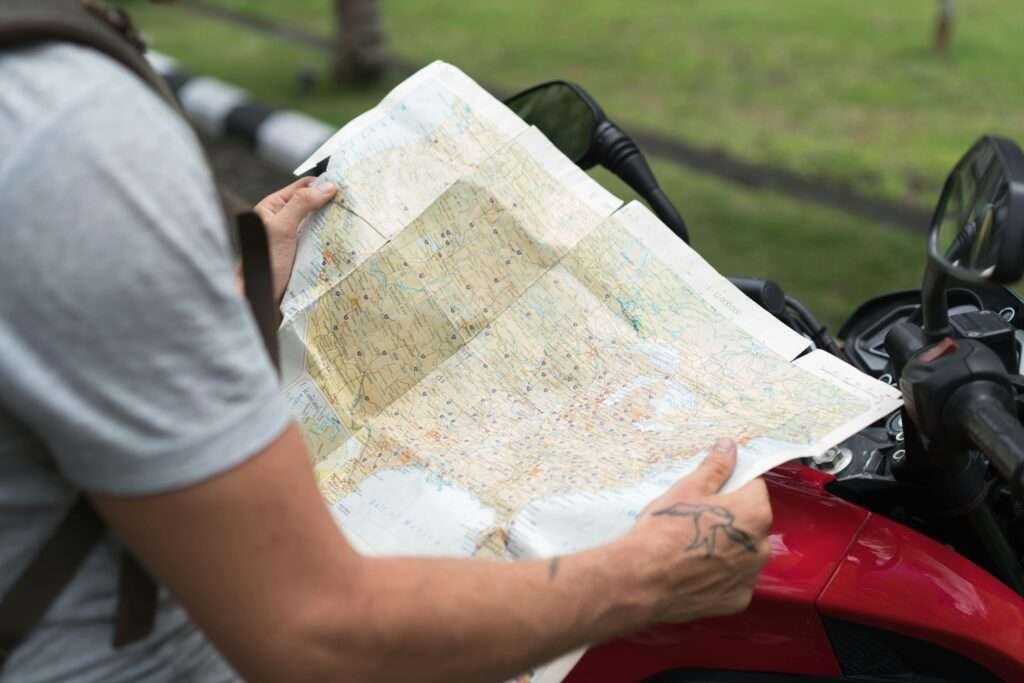 Side view of crop unrecognizable male in casual clothes with backpack sitting on motorcycle while studying city map on street